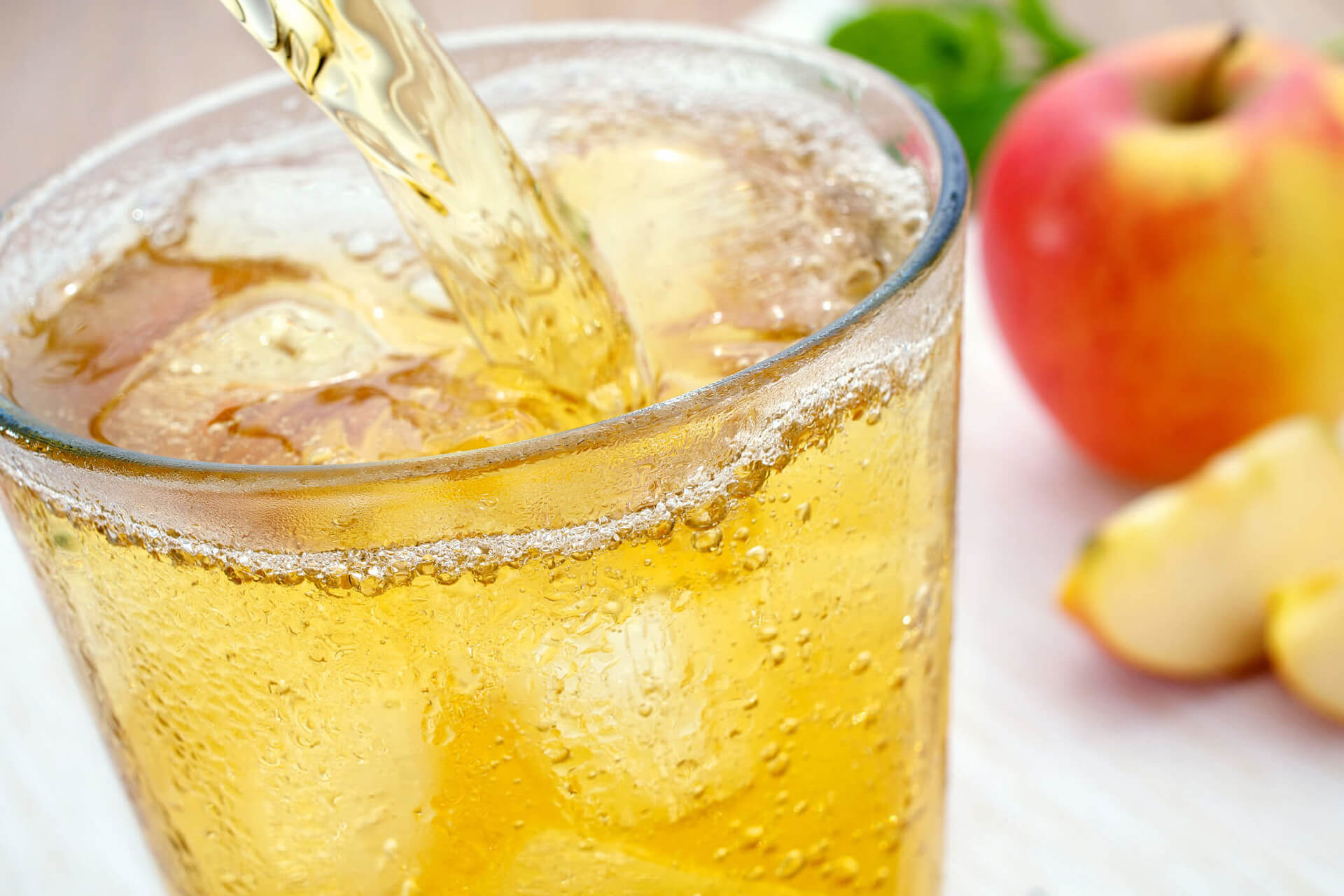In ein bereits gefülltes, eisgekühltes Glas mit Eiswürfeln wird Apfelschorle eingegossen. Im Hintergrund unscharf ein Apfel und zwei Apfelschnitze.