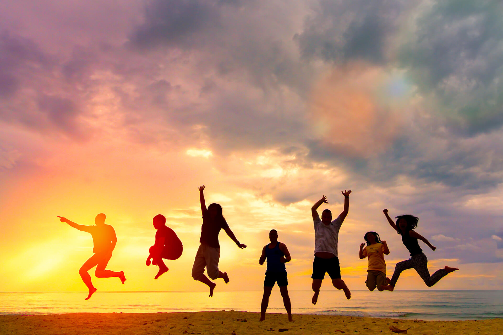 Sieben junge Menschen springen in jubelnder Pose an einem Strand wÃ¤hrend eines Sonnenuntergangs.